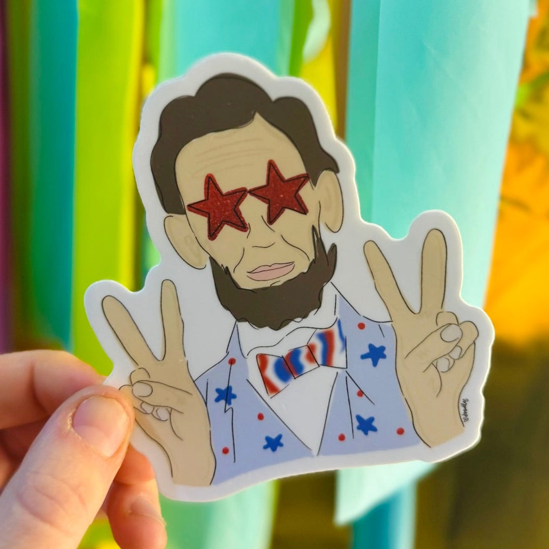 Doodles by Rebekah whimsical Abraham Lincoln sticker. This patriotic, pop-art style historical figure decal features red star glasses and a star-patterned suit, making it a unique and fun gift for history buffs to use on water bottles, laptops, and journals.