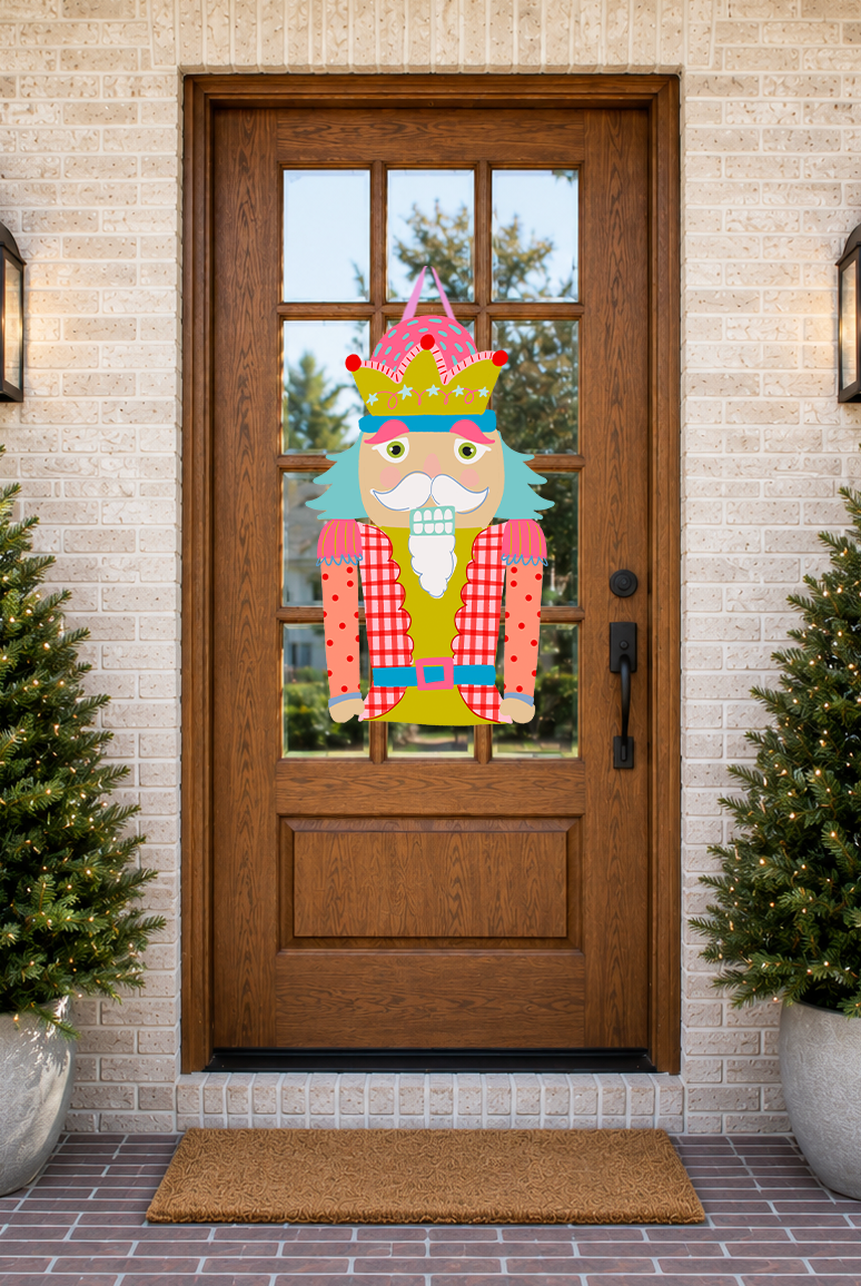 Decorative Nutcracker door hanger on a wooden front door with Christmas trees and a doormat.