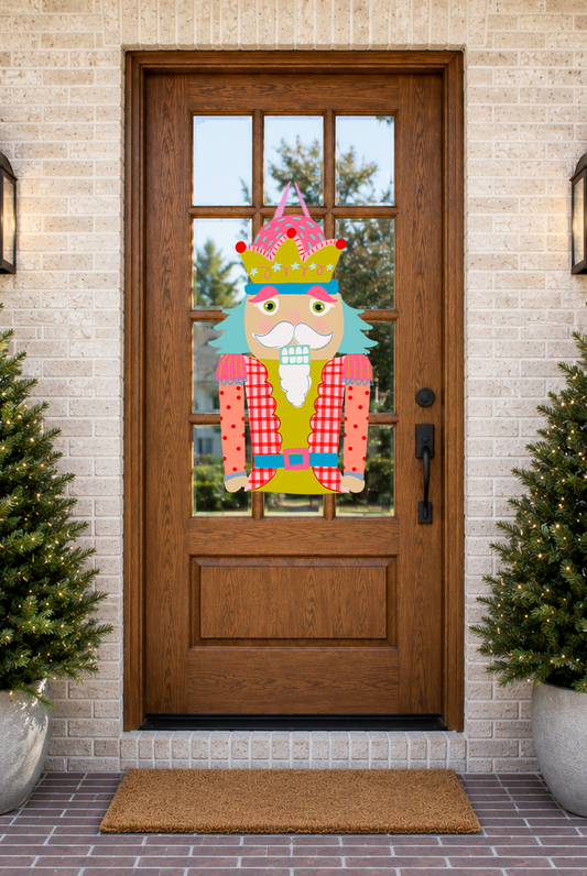 Decorative Nutcracker door hanger on a wooden front door with Christmas trees and a doormat.