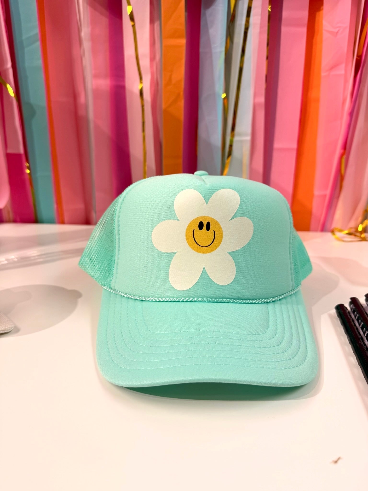 Mint green trucker hat with a whimsical white daisy smiley face graphic, featuring a hand-drawn floral aesthetic perfect for summer festivals or casual wear.