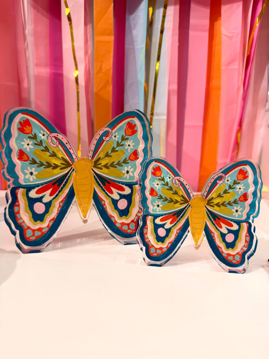 Doodles by Rebekah whimsical butterfly acrylic home decor featuring hand-painted floral wing designs in teal and red. These colorful spring butterfly accents are shown against a bright party streamer background.