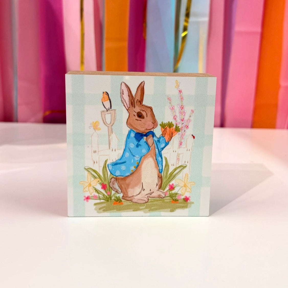 Doodles by Rebekah "Peter Rabbit" inspired wooden block home decor featuring a hand-painted bunny in a blue coat. This whimsical spring garden-themed tabletop or shelf accent is shown against a bright party streamer backdrop.
