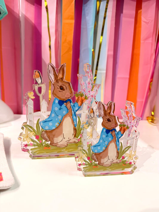 Doodles by Rebekah Easter bunny acrylic home decor featuring a whimsical hand-painted rabbit in a blue coat holding carrots. These colorful spring tabletop accents are shown against a bright party streamer background.