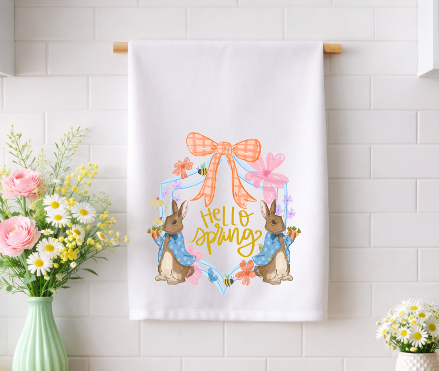 "Hello Spring" decorative kitchen tea towel with whimsical bunny and floral garden illustrations displayed against a white tile backsplash.