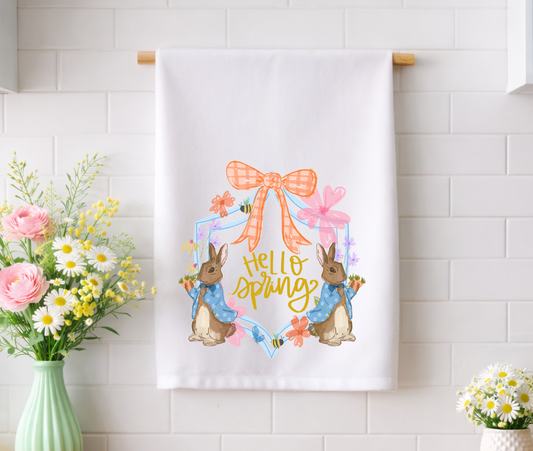 "Hello Spring" decorative kitchen tea towel with whimsical bunny and floral garden illustrations displayed against a white tile backsplash.