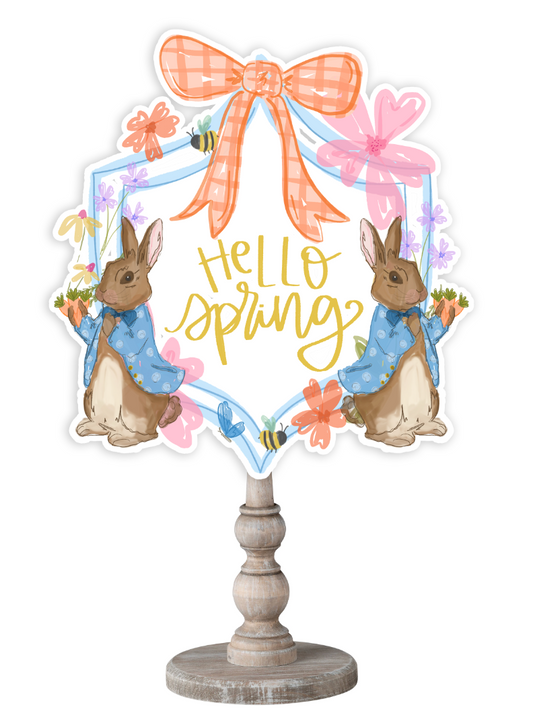 Doodles by Rebekah spring decor featuring a whimsical Peter Rabbit-style illustration, an orange gingham bow, and hand-lettered "Hello Spring" text on a rustic wood stand.