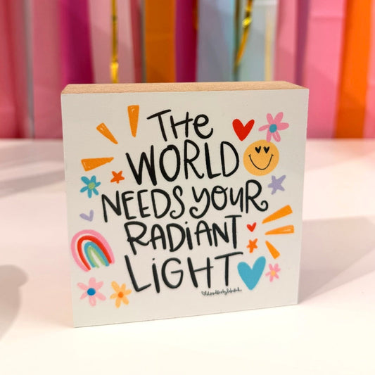 Doodles by Rebekah inspirational wooden block home decor featuring "The World Needs Your Radiant Light" whimsical hand-painted typography. This bright, positive tabletop or shelf accent is shown against a colorful party streamer background.