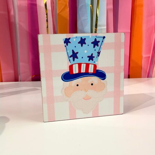 Doodles by Rebekah patriotic Uncle Sam wooden block home decor featuring a whimsical hand-painted design on a pink checkered pattern. This Americana-themed tabletop or shelf accent is shown against a bright party streamer background.