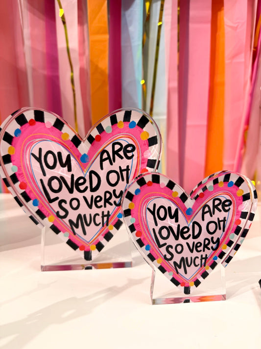 Doodles by Rebekah "You Are Loved" heart acrylic home decor featuring whimsical hand-painted checkered borders and multi-colored dots. These colorful Valentine's Day or nursery tabletop accents are shown against a bright party streamer background.